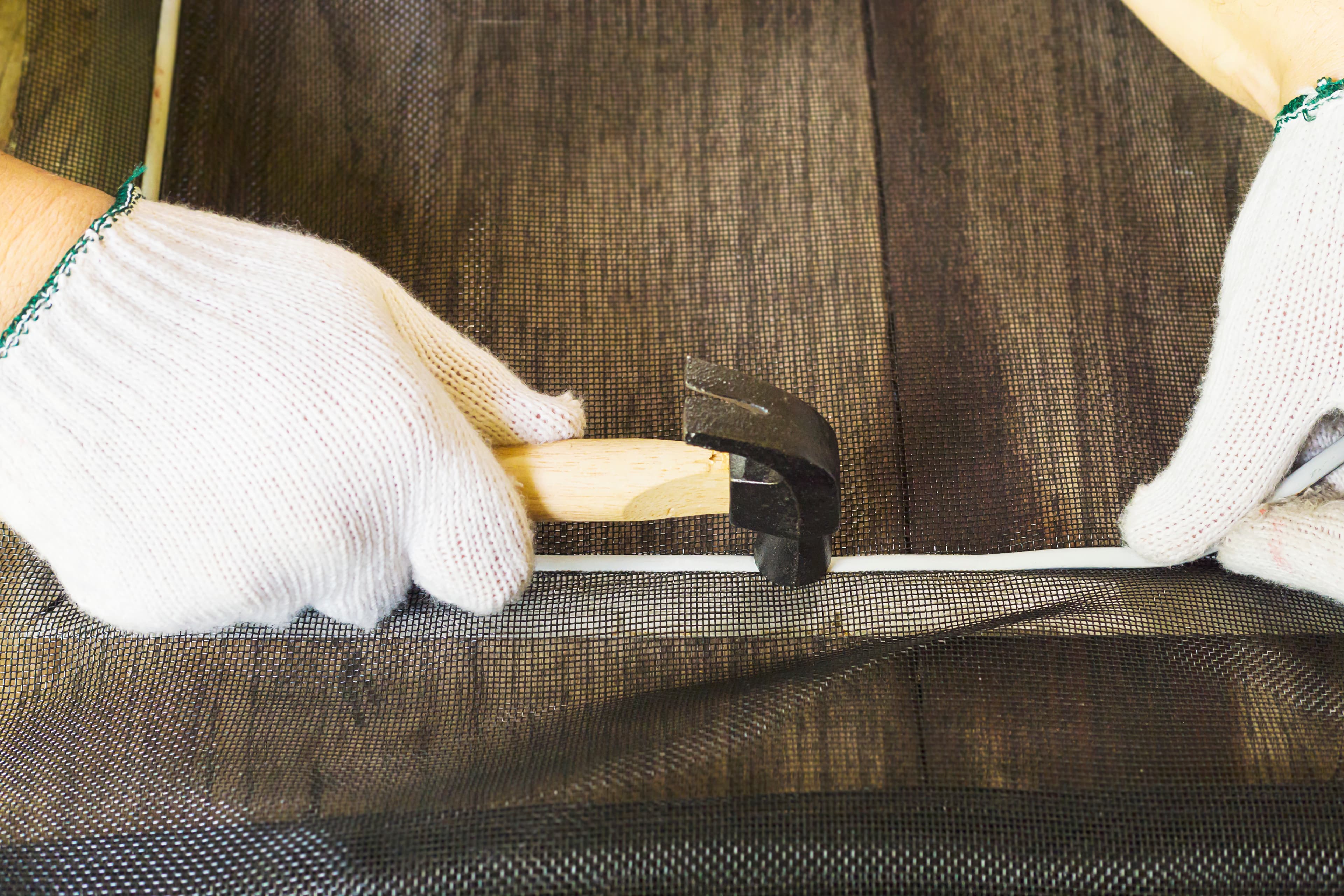 Hände mit Arbeitshandschuhen befestigen ein Gewebe mit einem Hammer an einem Rahmen.